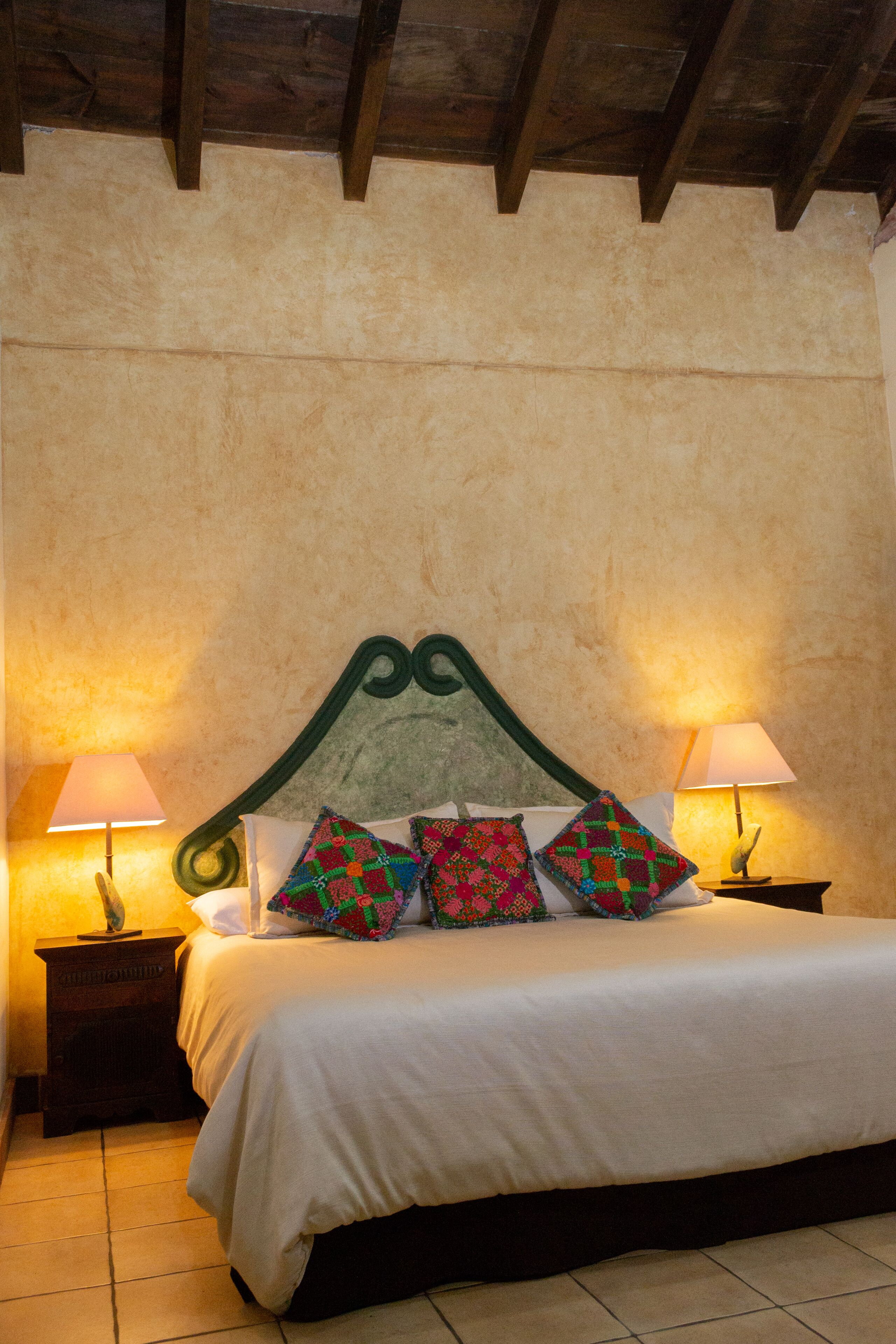 Una cama impecablemente arreglada con cabecera verde, dos lámparas de noche y almohadas coloridas.