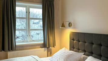 Une chambre avec une fenêtre, un radiateur, un lit avec une literie blanche, une tête de lit et une lampe de chevet.