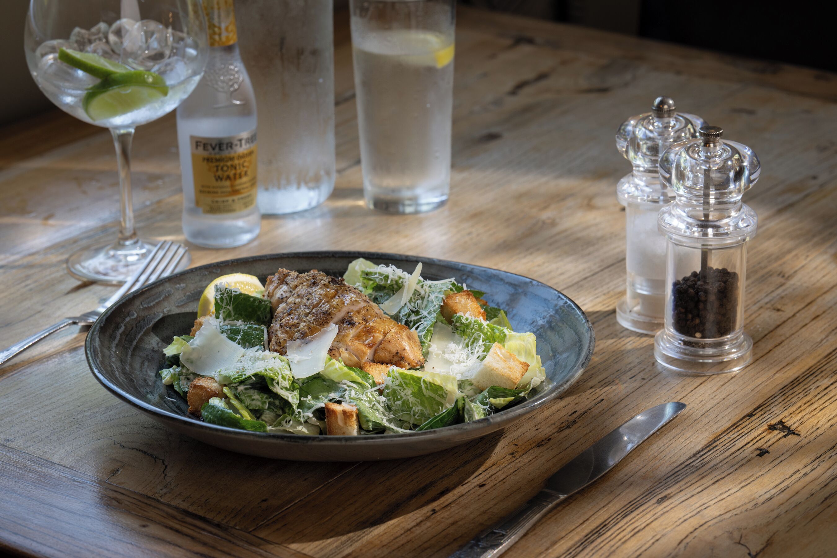 A table with a plate of food, a bottle of tonic water, and two salt and pepper shakers.