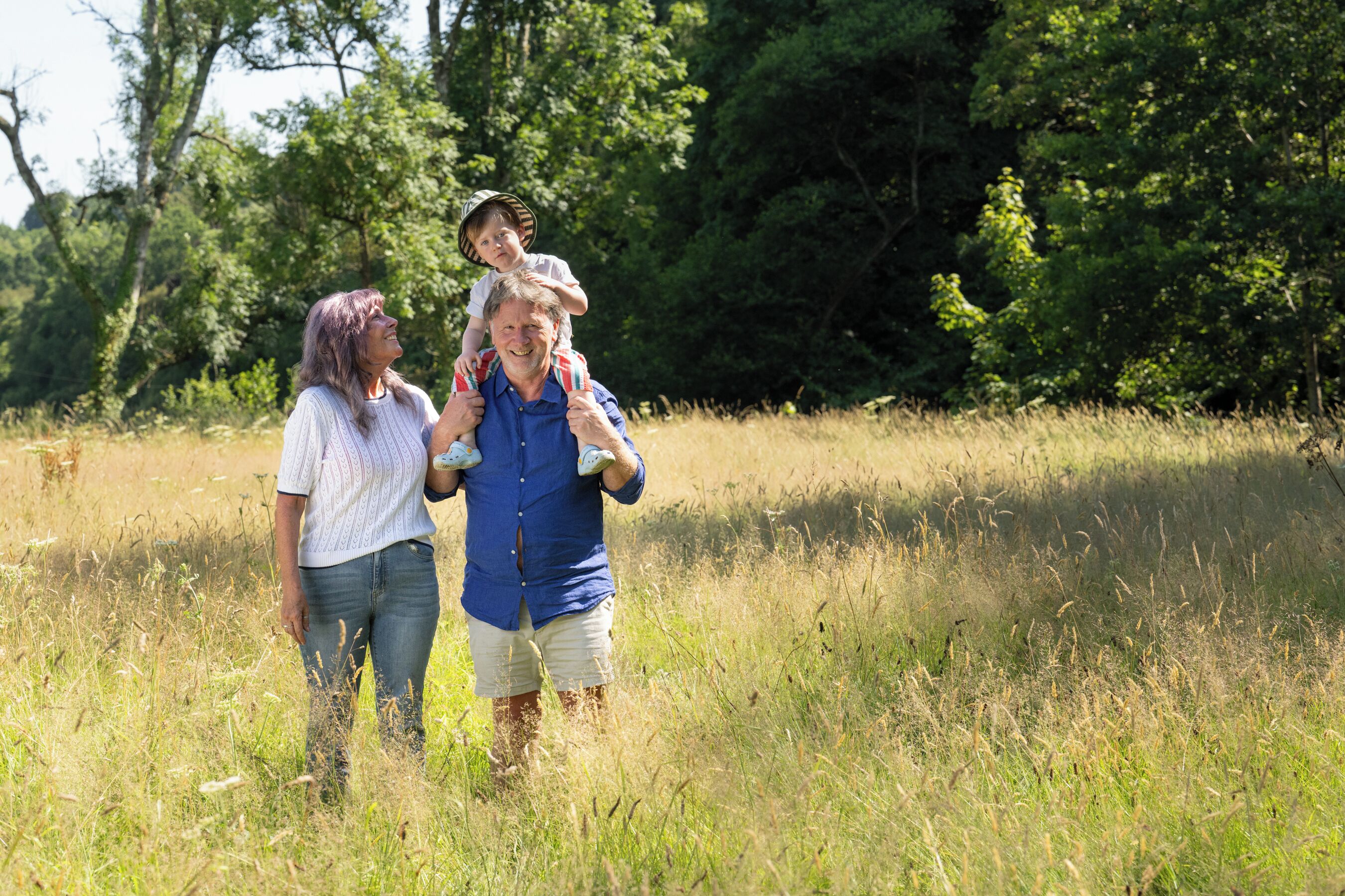 A person carrying a child on their shoulders in a grassy field with trees in the background.