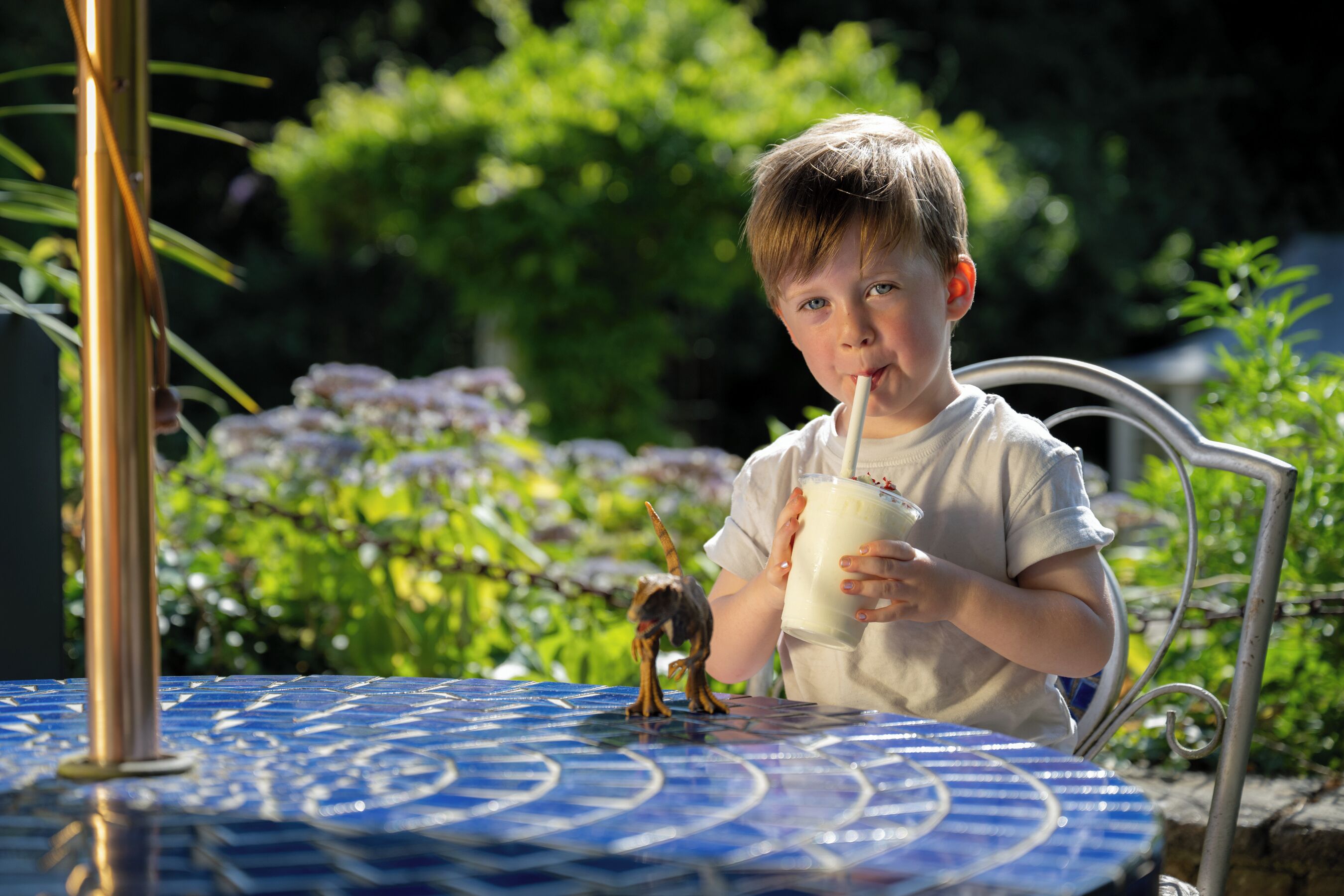A child sitting at a table with a drink, outdoors with greenery in the background.