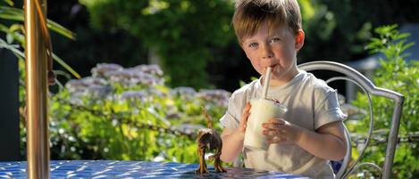 A child sitting at a table with a drink, outdoors with greenery in the background.