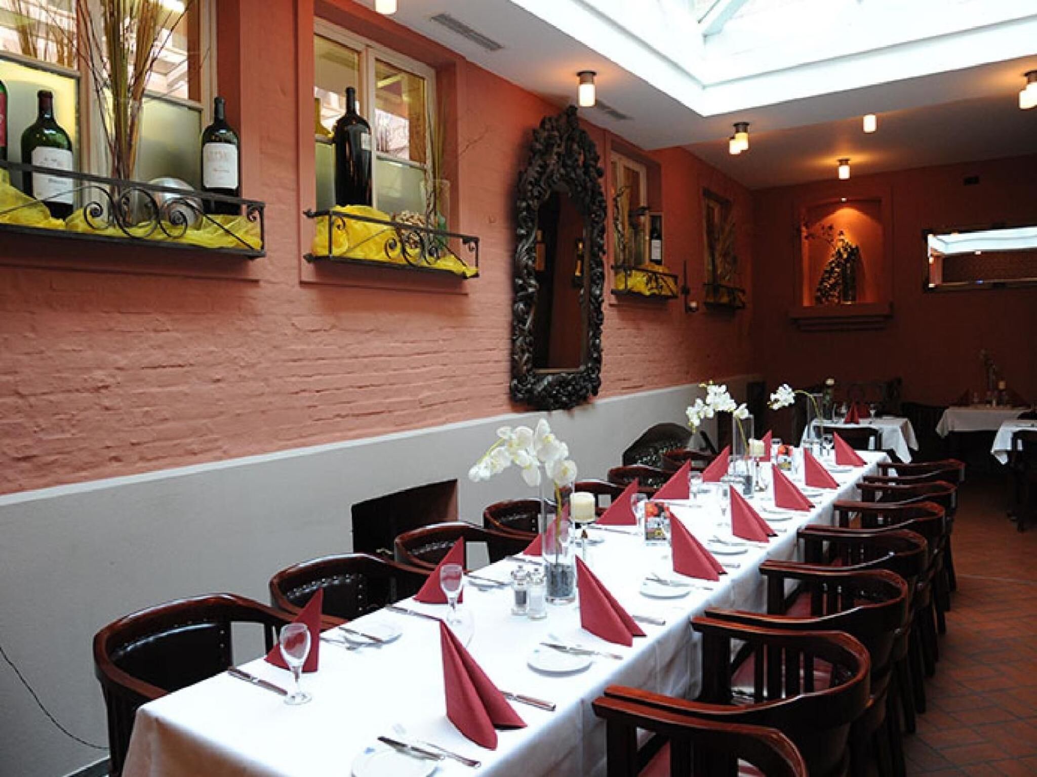 A restaurant with long tables set for dining, a brick wall, and a window with bottles.