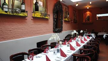 A restaurant with long tables set for dining, a brick wall, and a window with bottles.