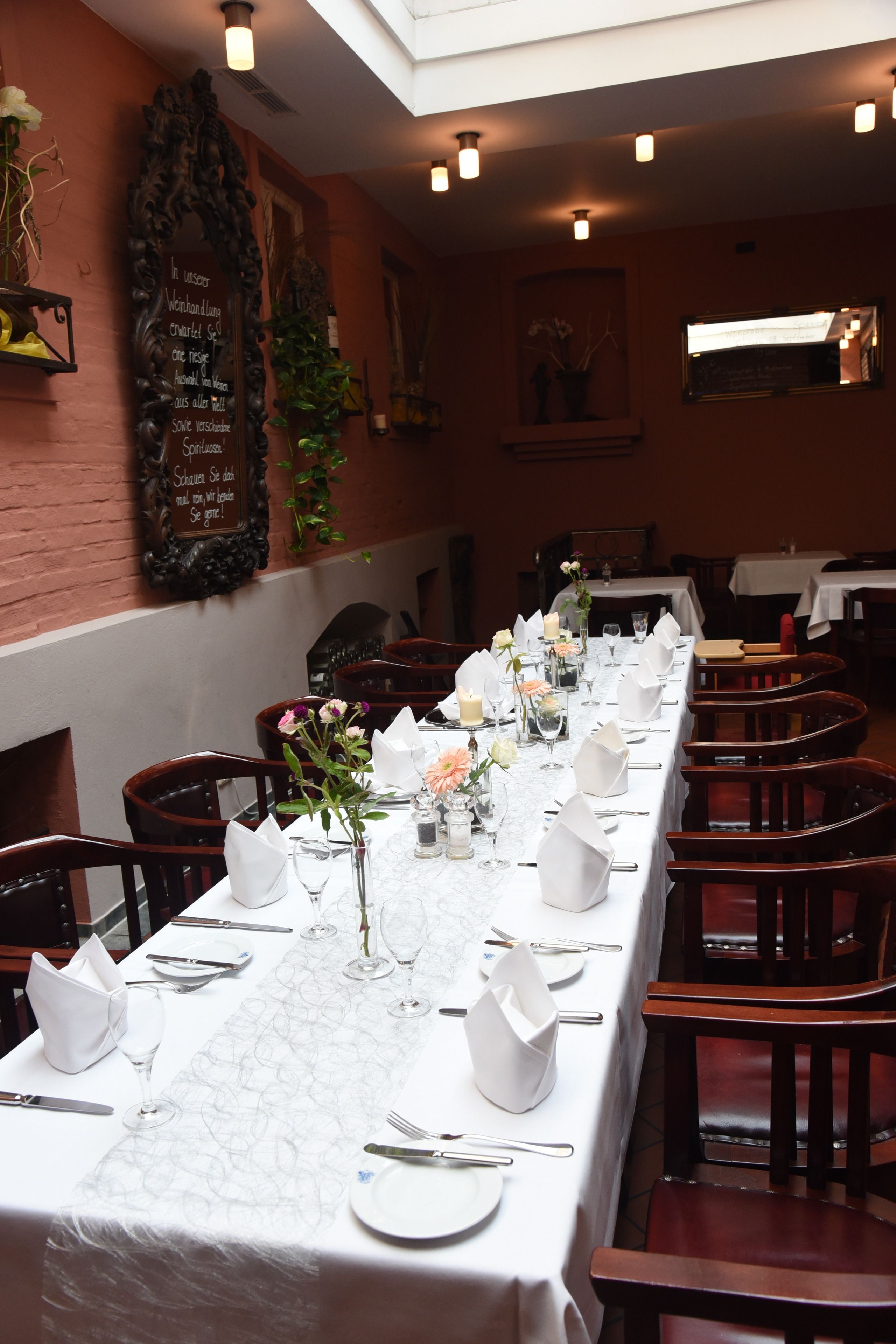 Et langt spisebord dækket med hvide dug, vinglas og blomsterarrangementer i en restaurant med murstensvægge og træstole.