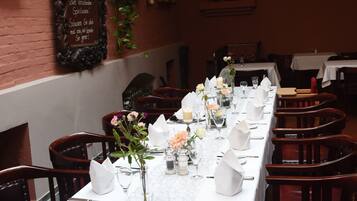 Et langt spisebord dækket med hvide dug, vinglas og blomsterarrangementer i en restaurant med murstensvægge og træstole.