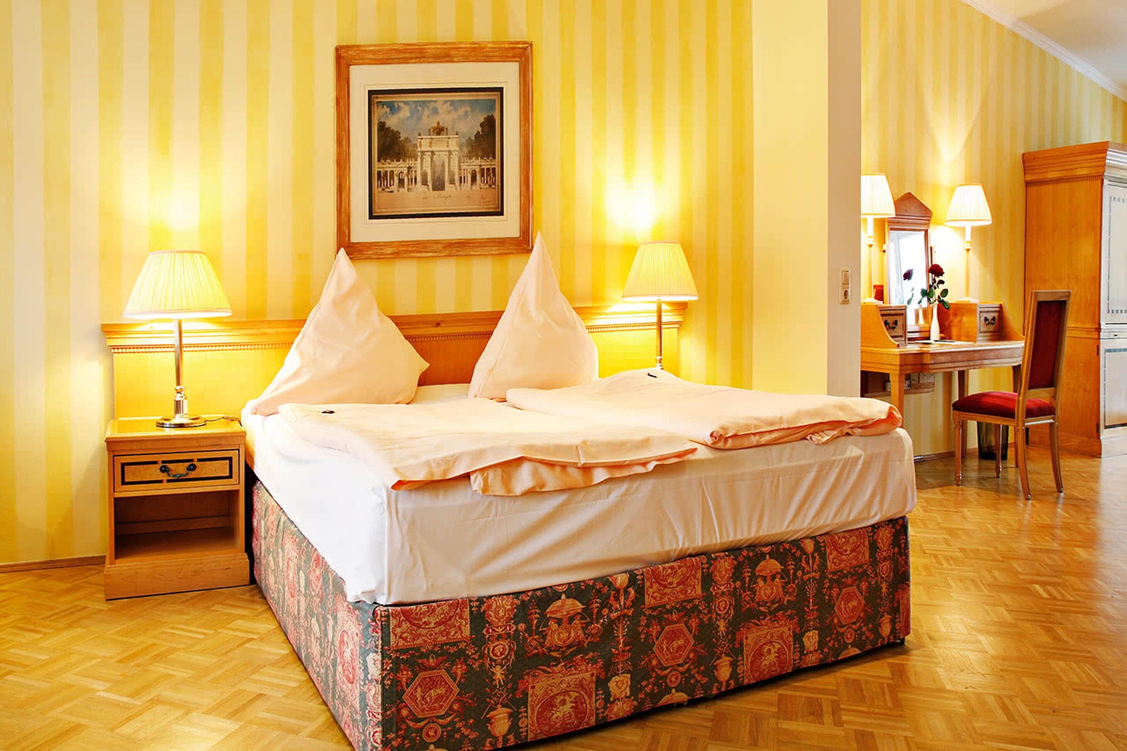 A hotel room with a large bed, two bedside tables with lamps, a wooden nightstand, a floral-patterned bench, a desk with a chair, and a framed picture on the wall.