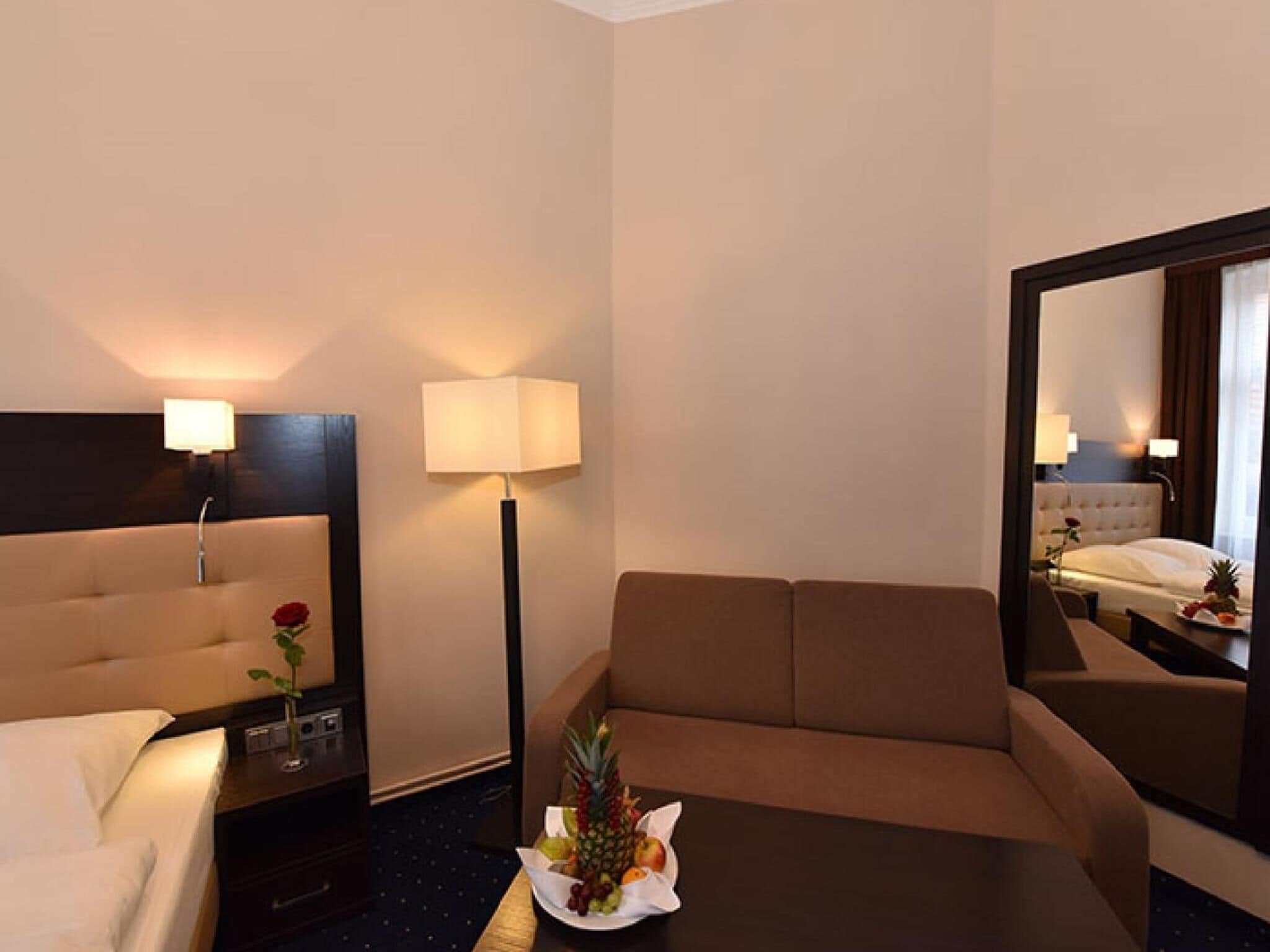 Una habitación de hotel con cama, sofá, mesa con fruta y un florero, lámpara y espejo.