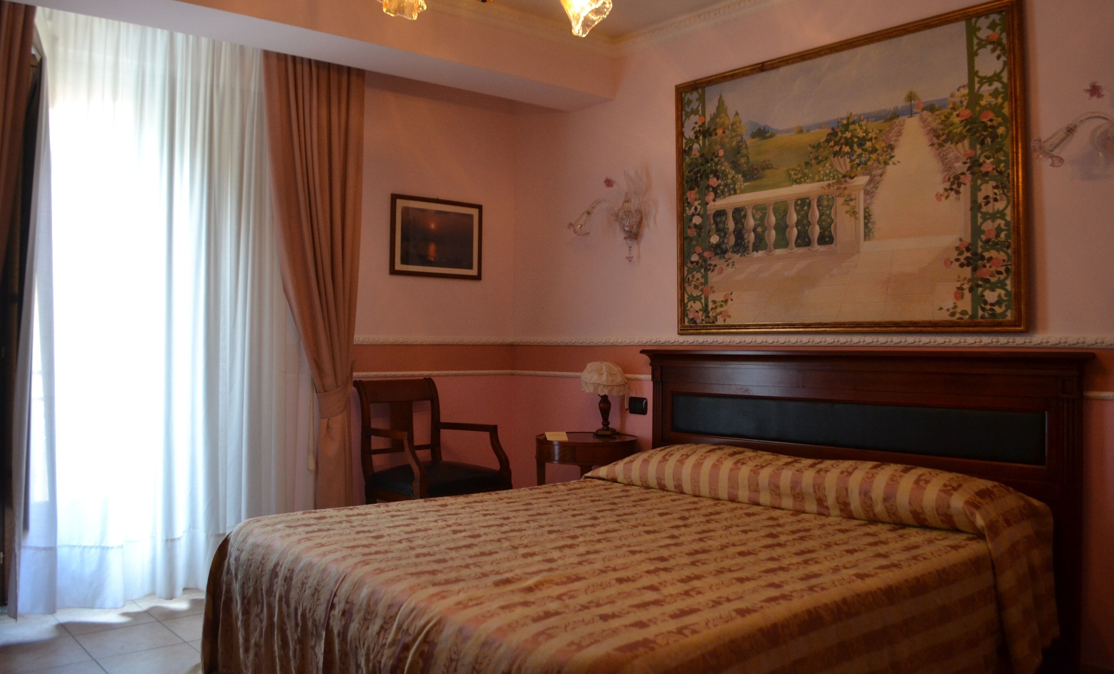 A hotel room with a bed, a chair, a painting, and a window with curtains.