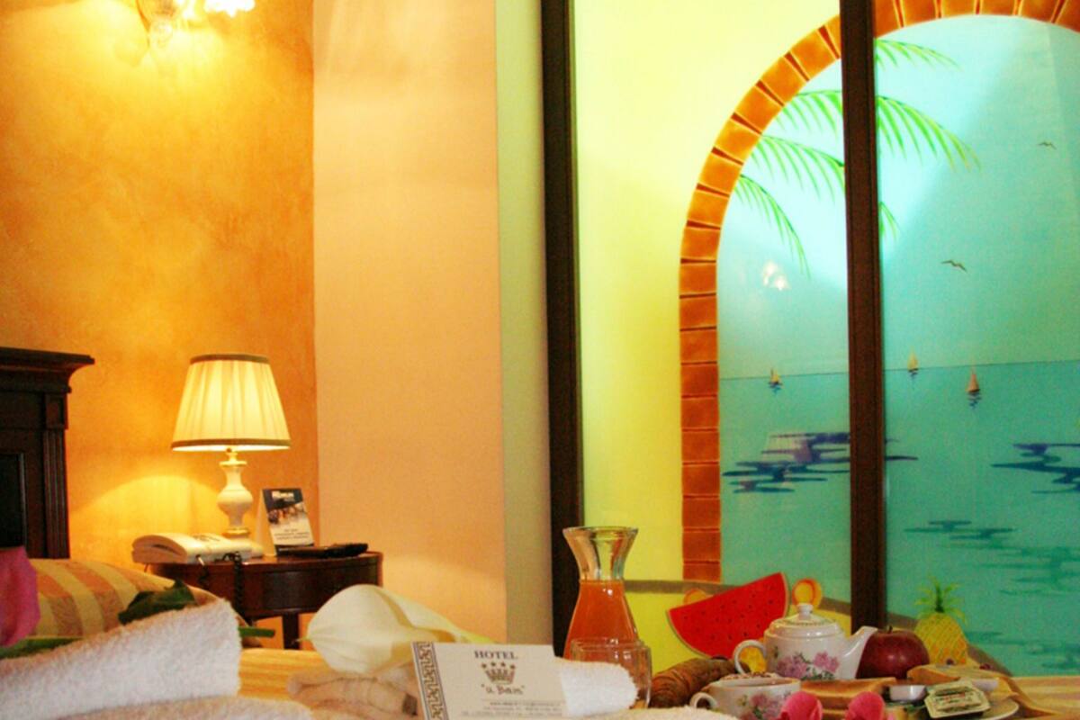 A hotel room with a bed, a nightstand, a lamp, a view of the sea, and a tropical-themed window.