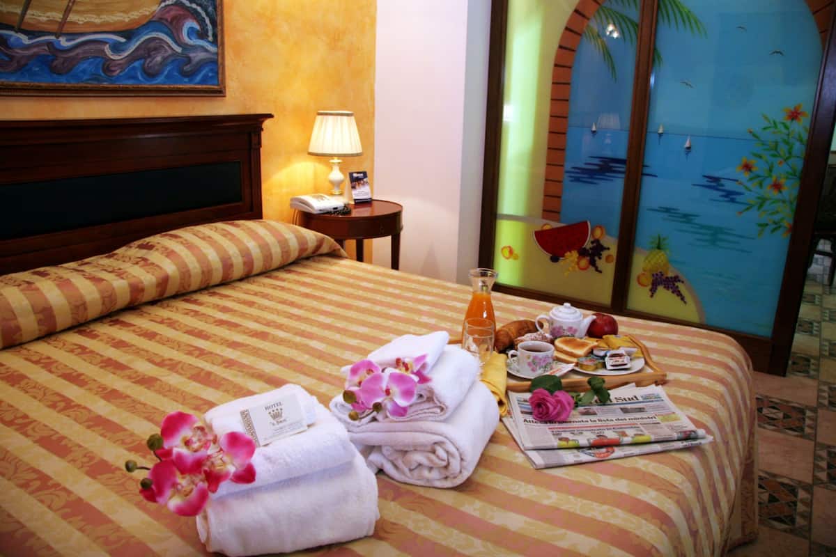A hotel room with a bed, a nightstand, a lamp, a window with a view of palm trees, and a breakfast tray on the bed.