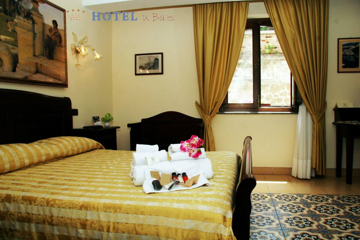 A hotel room with a bed, a nightstand, a window with curtains, and a painting on the wall.