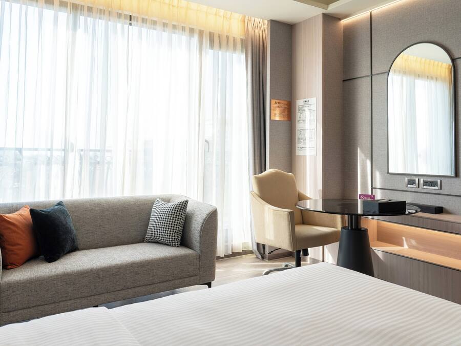 A modern hotel room with a bed, a sofa, a chair, a small table, and a large mirror.