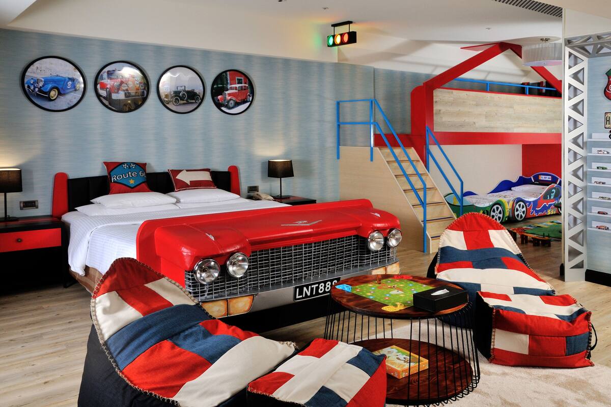A room with a bed, a red car-shaped headboard, a bunk bed, a desk, and a chair with a red, white, and blue cushion.