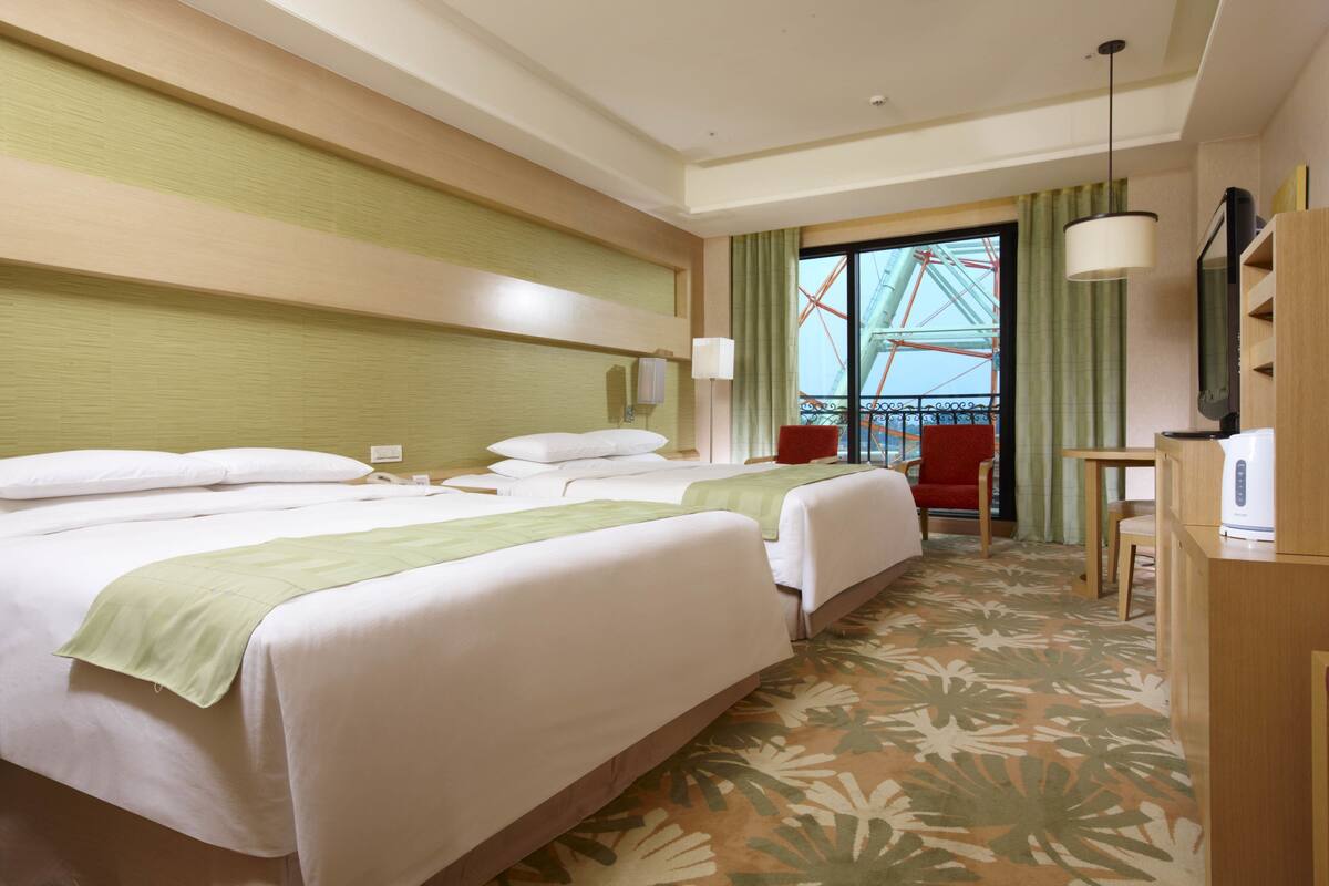 A hotel room with two beds, a desk, a chair, a TV, and a view of a Ferris wheel.