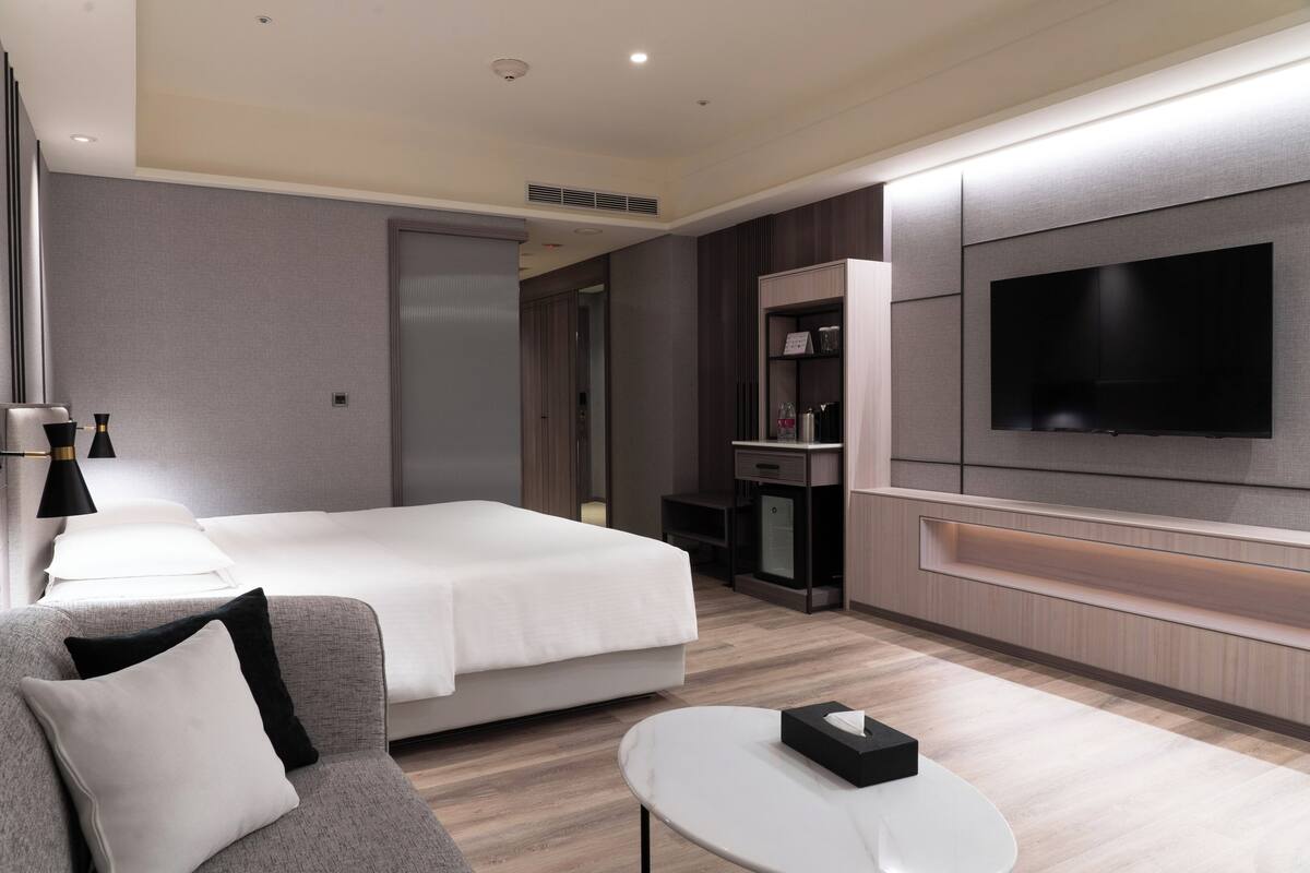 A modern hotel room with a large bed, a sofa, a TV, and a small desk.