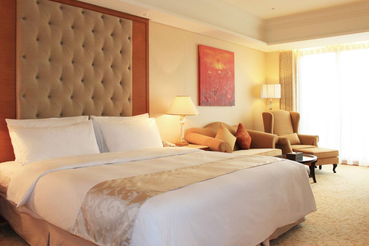 A hotel room with a large bed, a tufted headboard, a sofa, a chair, and a painting on the wall.