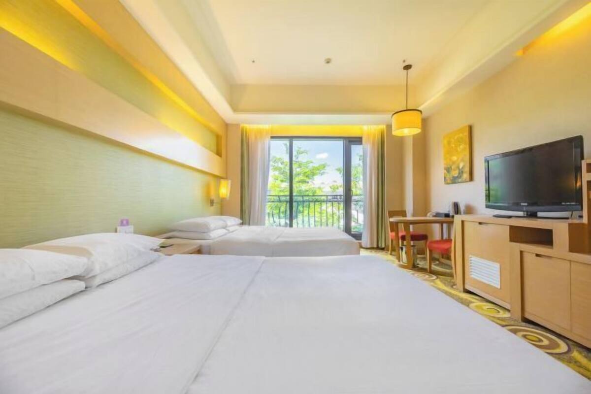 A hotel room with two beds, a TV, a desk, and a balcony with a view of greenery.