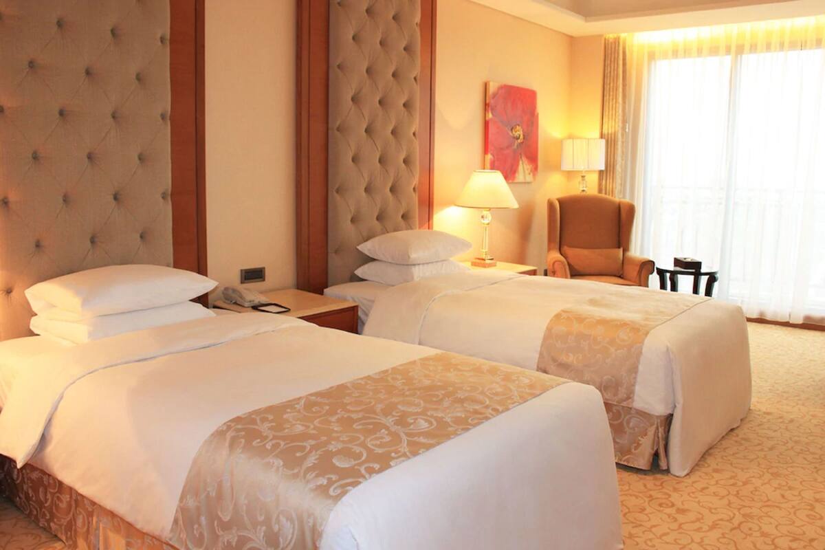 A hotel room with two beds, a sitting area with a chair, a lamp, and a painting on the wall.