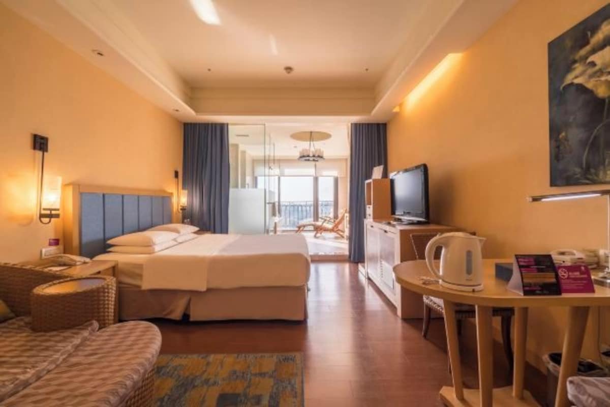 A hotel room with a bed, a desk with a kettle, a television, and a view of the balcony.