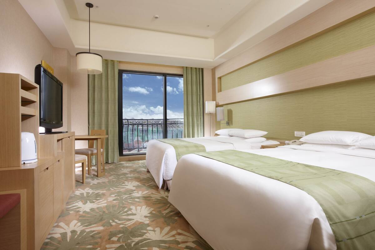A hotel room with two beds, a TV, a desk, and a balcony with a view.