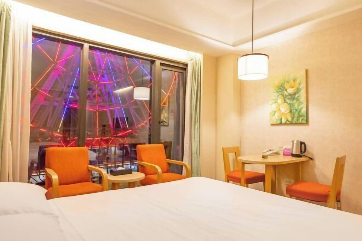A hotel room with a bed, orange chairs, a small table, a painting, and a view of a Ferris wheel.