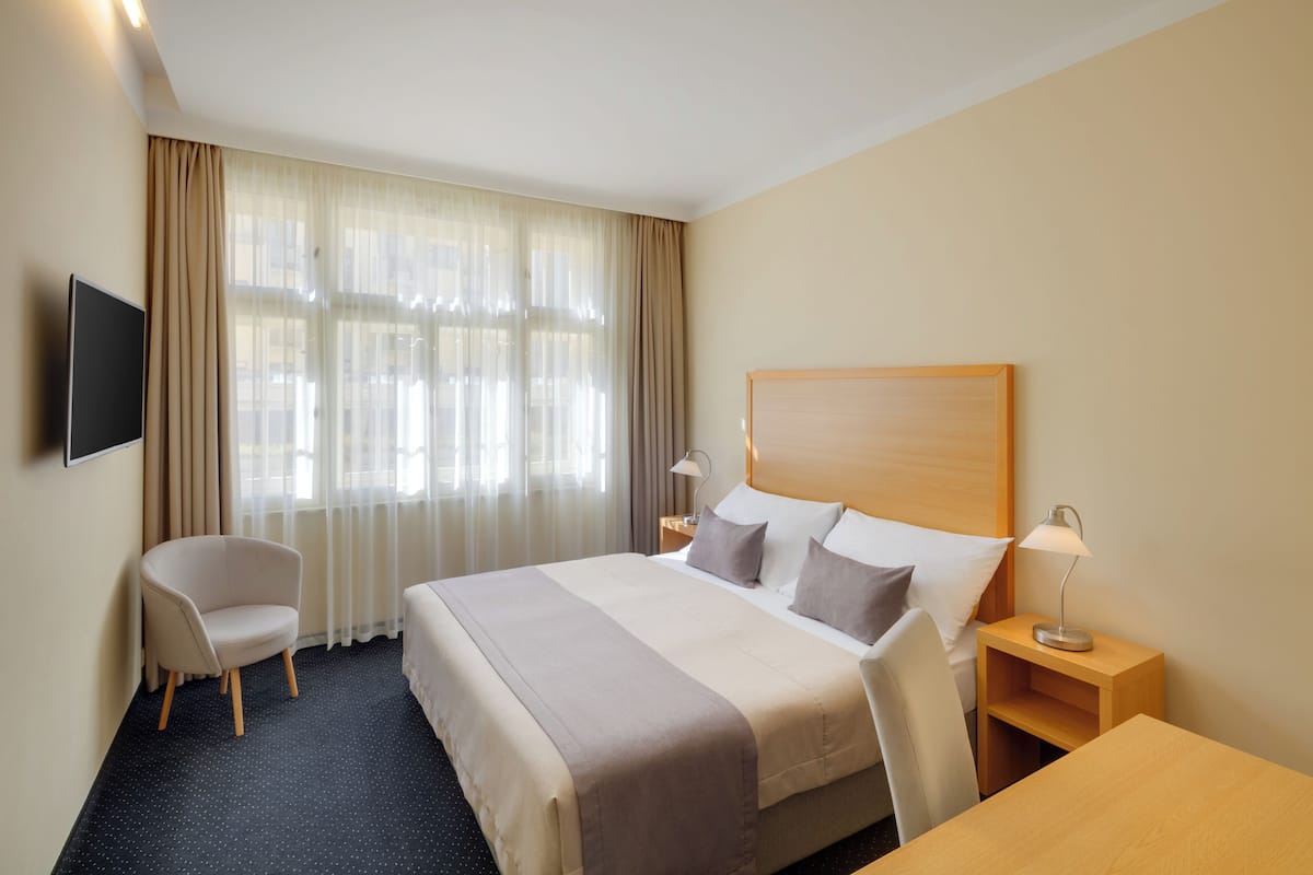 Habitación de hotel con una cama grande, una silla, un escritorio, una mesita de noche con lámpara y un televisor montado en la pared.