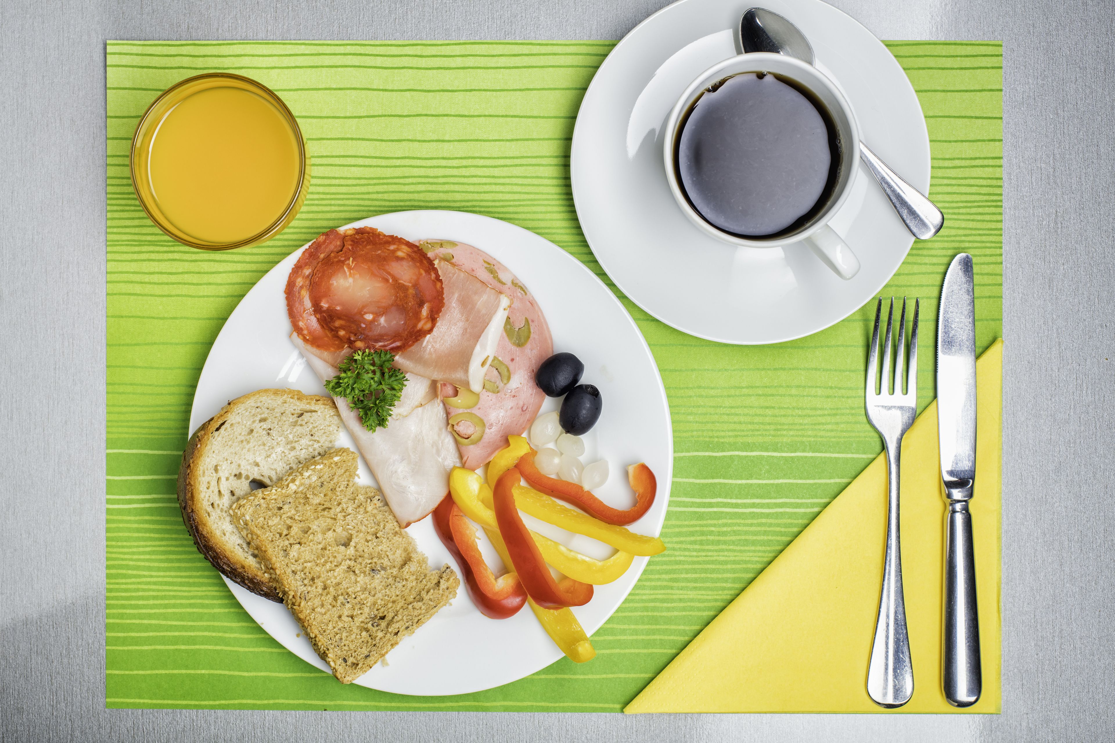 Une assiette de petit-déjeuner avec du jambon, des œufs, du pain et des légumes, une tasse de café et un verre de jus d’orange, le tout posé sur un set de table vert.