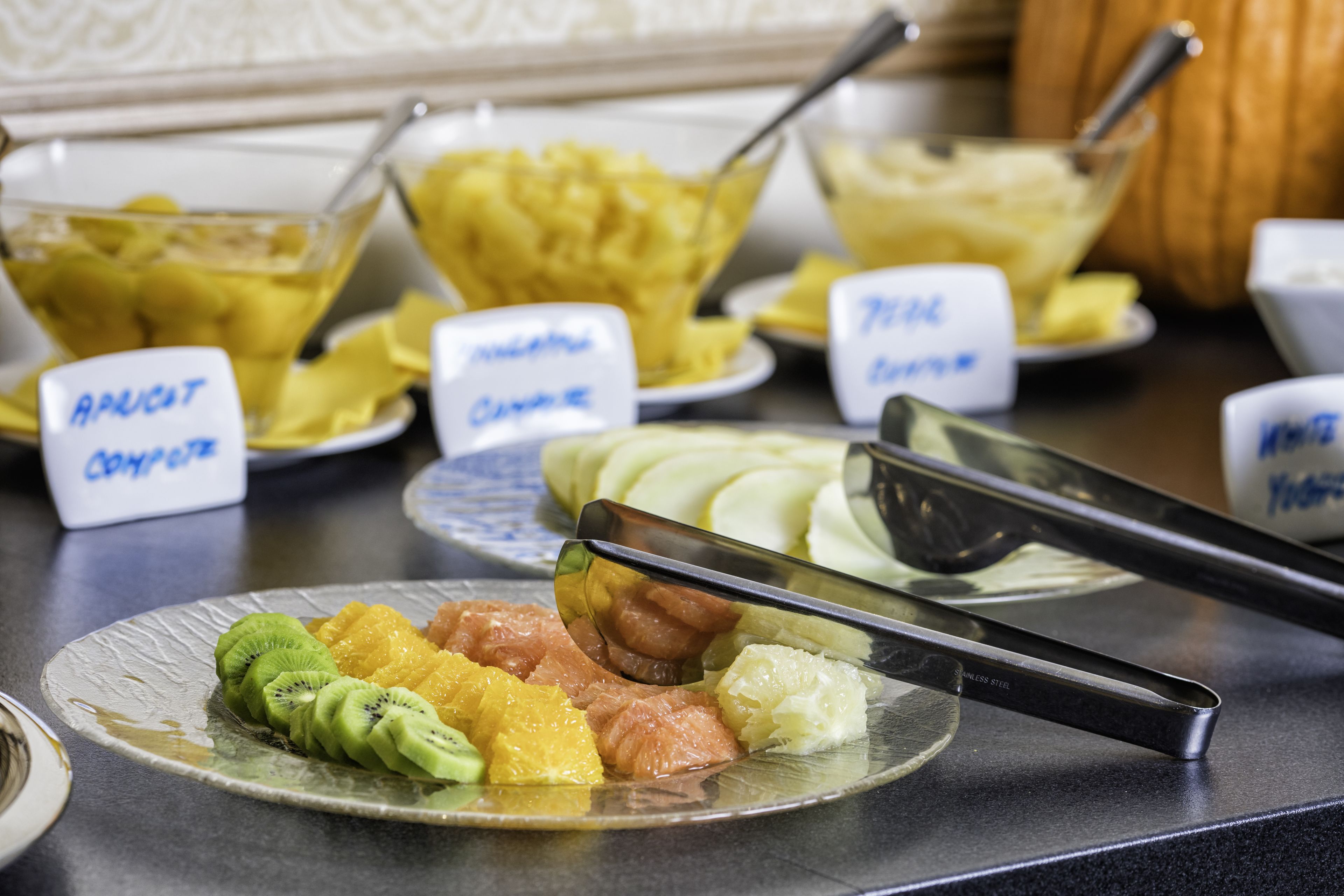 Un bufé con platos identificados, que incluye frutas y postres.