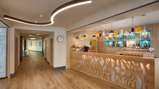 A modern hotel lobby with a wooden reception desk, a clock, and a bar area with shelves of bottles. - Hotel Clement (Prague)
