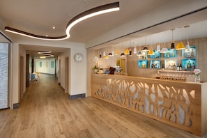 A modern hotel lobby with a wooden reception desk, a clock, and a bar area with shelves of bottles. - Hotel Clement (Prague)