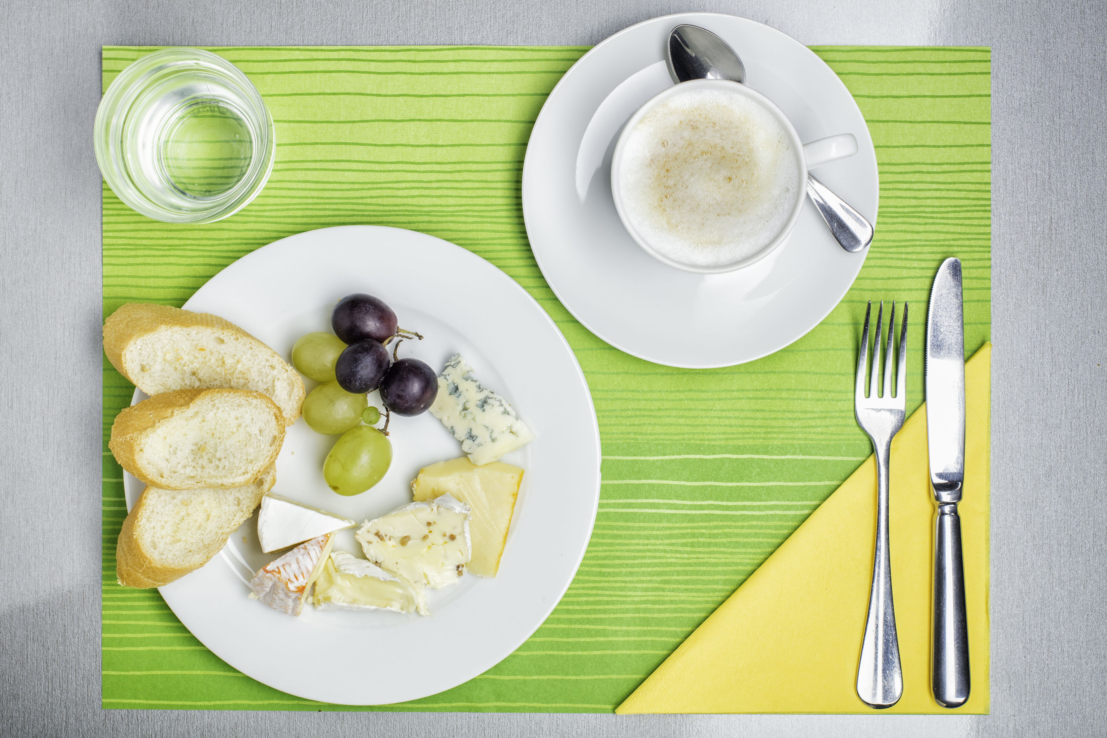 Un desayuno con un plato de queso, uvas y pan, una taza de café y cubiertos sobre una servilleta verde.