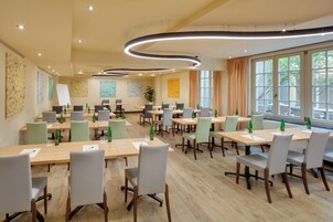 Uma sala de conferência com várias mesas e cadeiras, equipada com garrafas de água e copos, e grandes janelas que proporcionam luz natural.