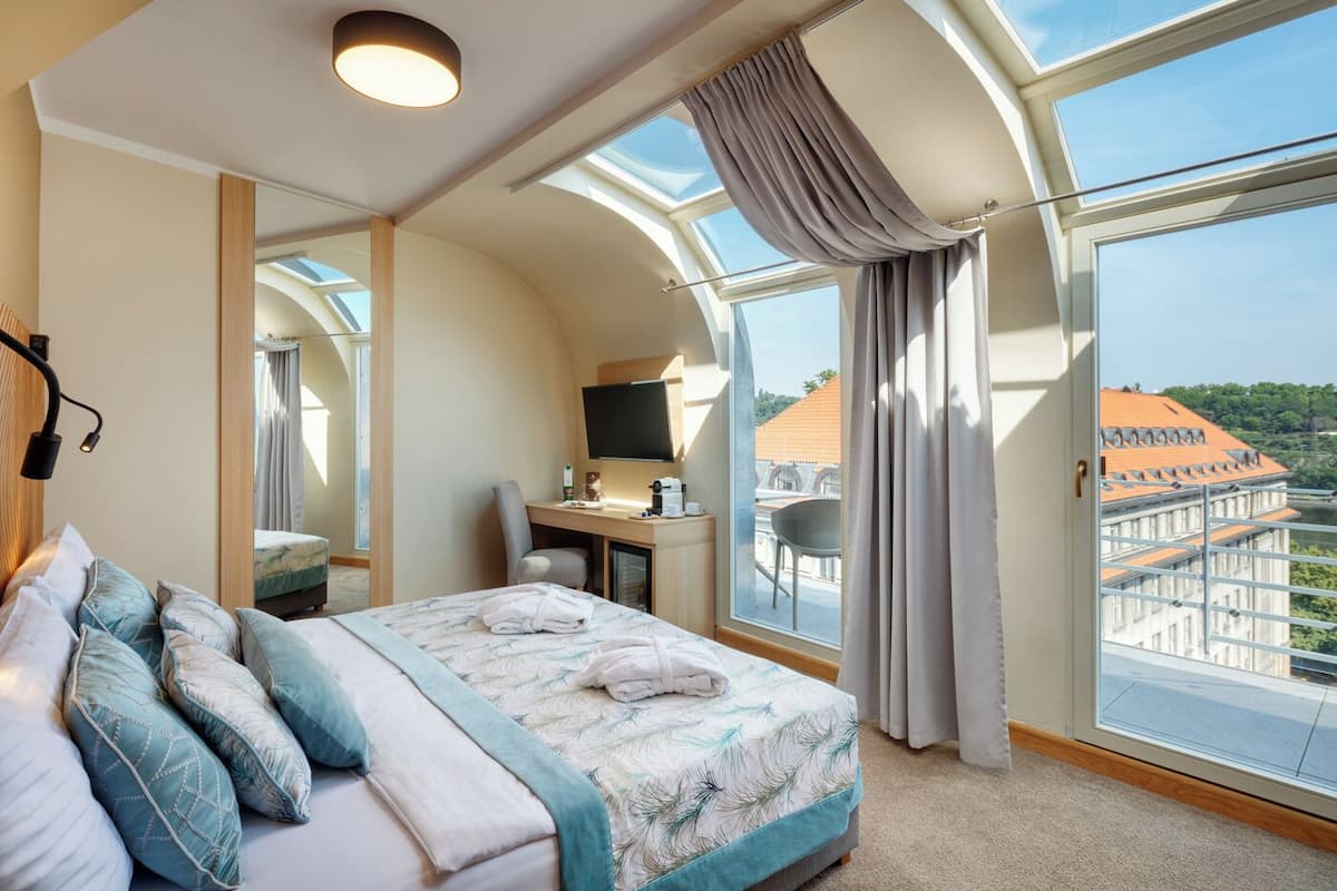 Habitación de hotel moderna con una cama grande, un escritorio, una silla, un televisor y un balcón con vistas a la ciudad.