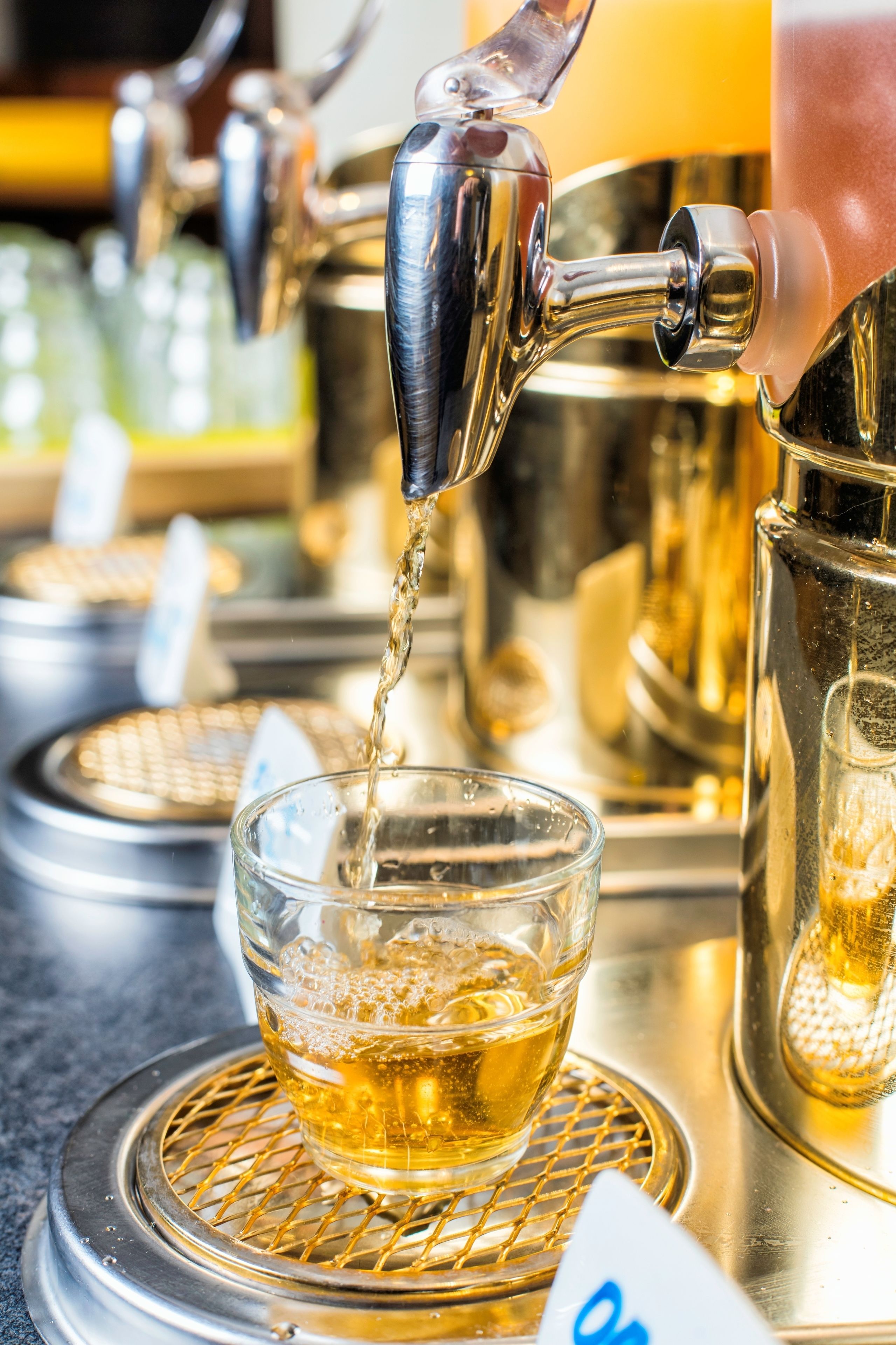 A hand pouring a golden liquid from a tap into a glass on a metal tray.