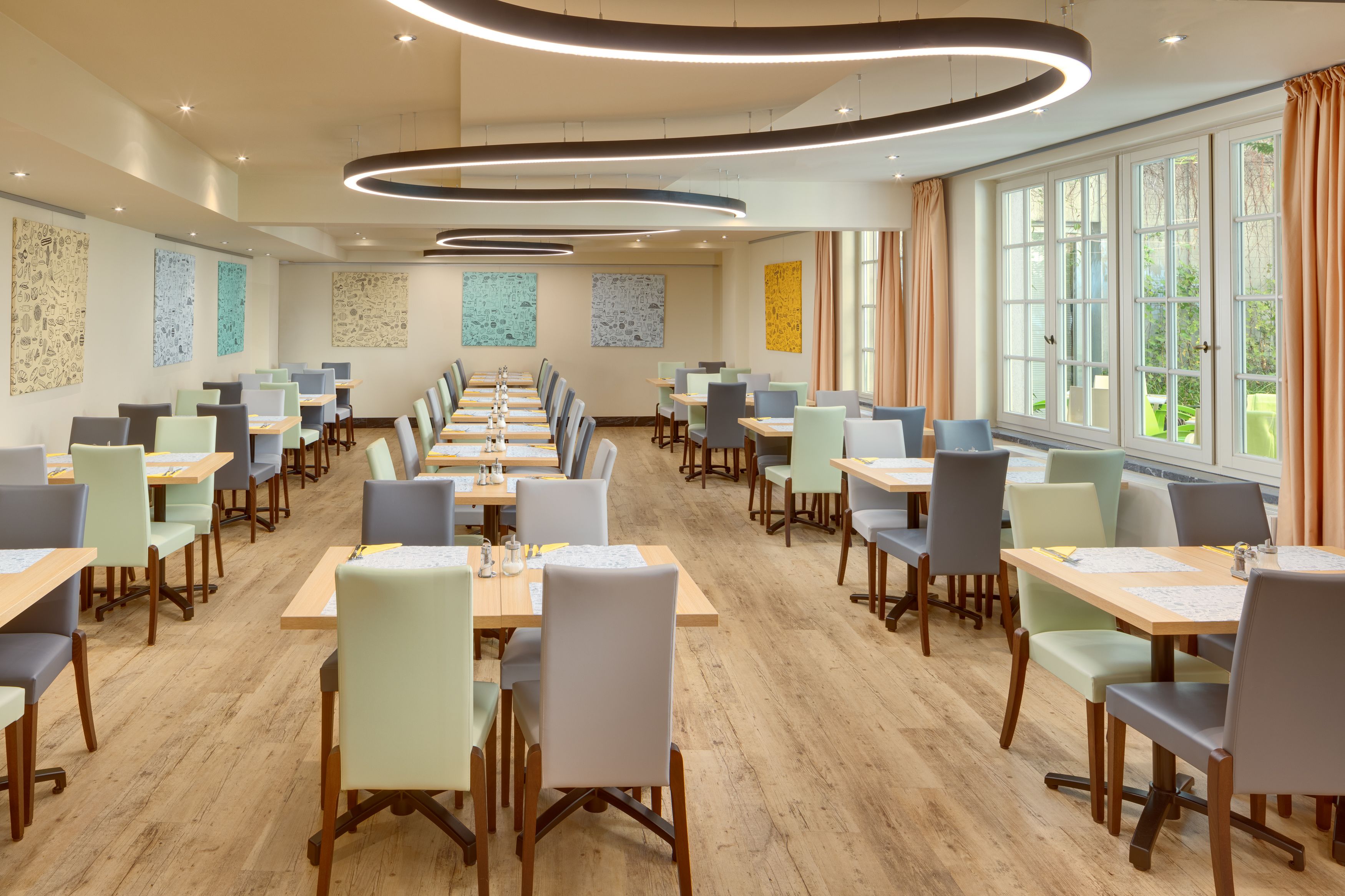 A modern dining area with long tables and chairs, large windows, and abstract wall art.