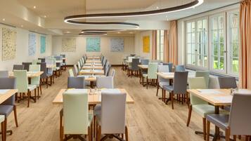 A modern dining area with long tables and chairs, large windows, and abstract wall art.