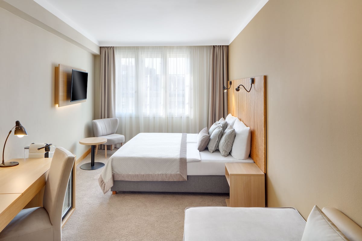 Habitación de hotel con cama, escritorio, silla, televisor y ventana con cortinas.