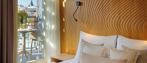 A hotel room with a large bed, a wooden headboard, a small table, and a view of the city through a window.