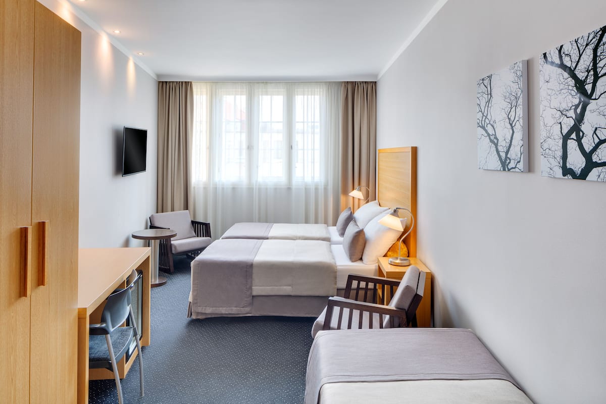 Una habitación de hotel con cama, escritorio, silla, mesita, televisor y ventana con cortinas.