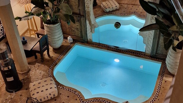 Une petite piscine intérieure avec un sol carrelé et une arche décorative. On y trouve deux chaises longues et une table recouverte d’une nappe à motifs.