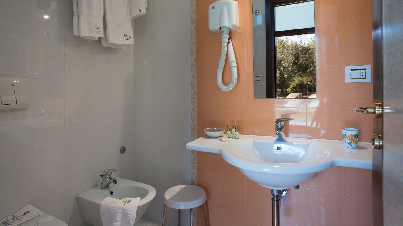 Ein Badezimmer mit einem weißen Waschbecken, einem Bidet und einer Toilette. Es gibt einen Handtuchhalter mit Handtüchern, ein Fenster mit Blick auf Bäume und eine Tür mit Griff.
