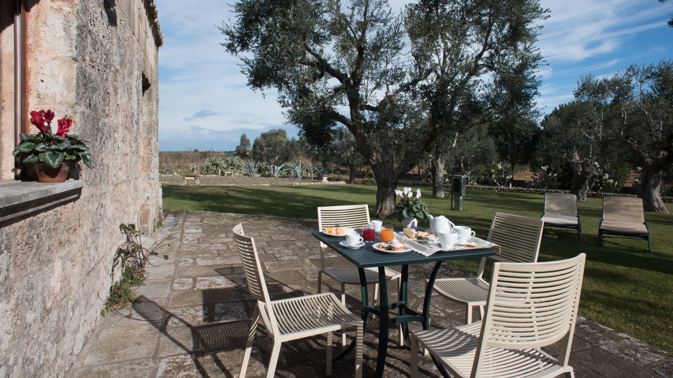 An outdoor dining area with a stone table and chairs, surrounded by trees and a stone wall.