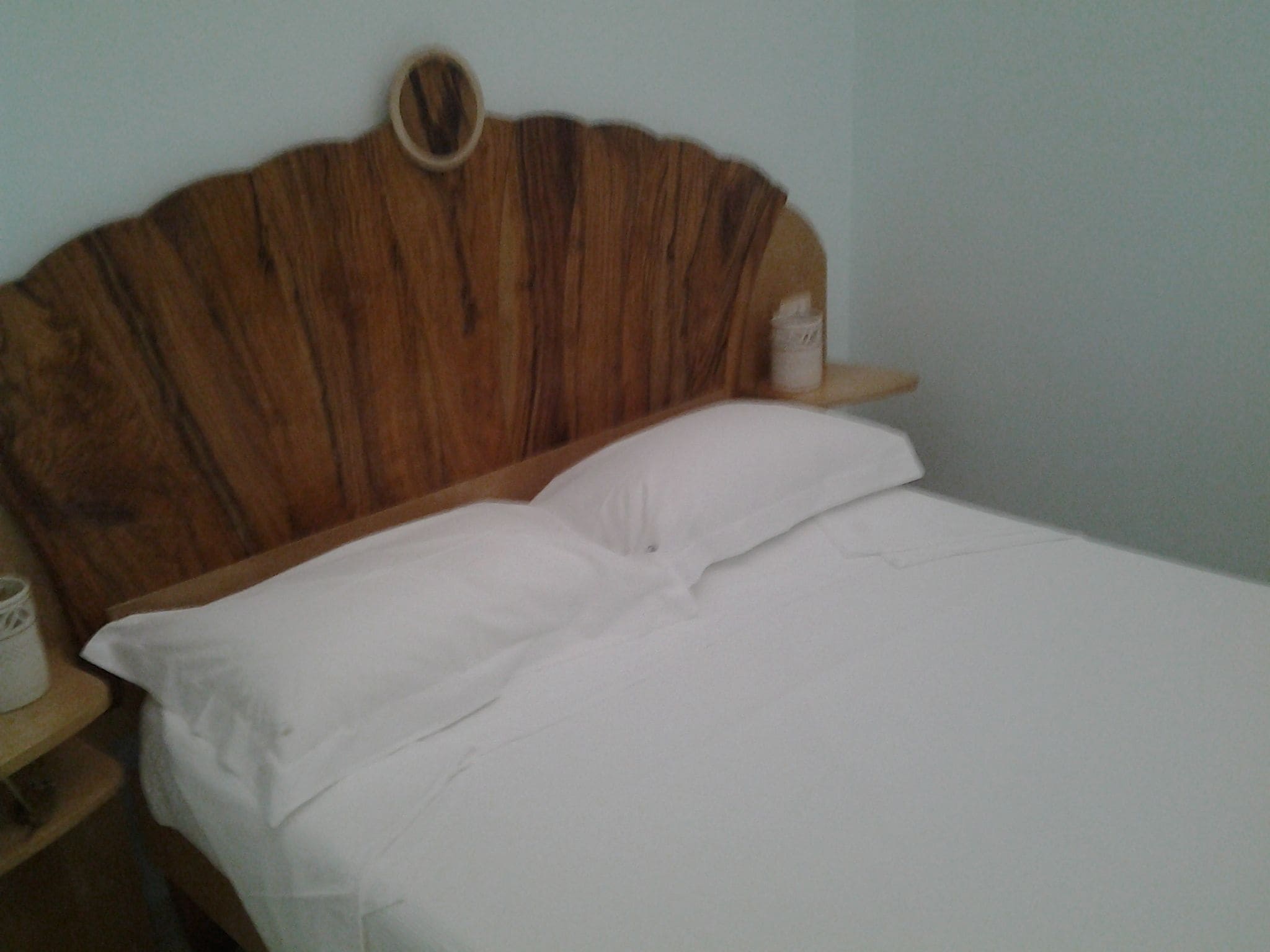 Een bed met witte lakens, een houten hoofdbord en een nachtkastje met een wit voorwerp erop.