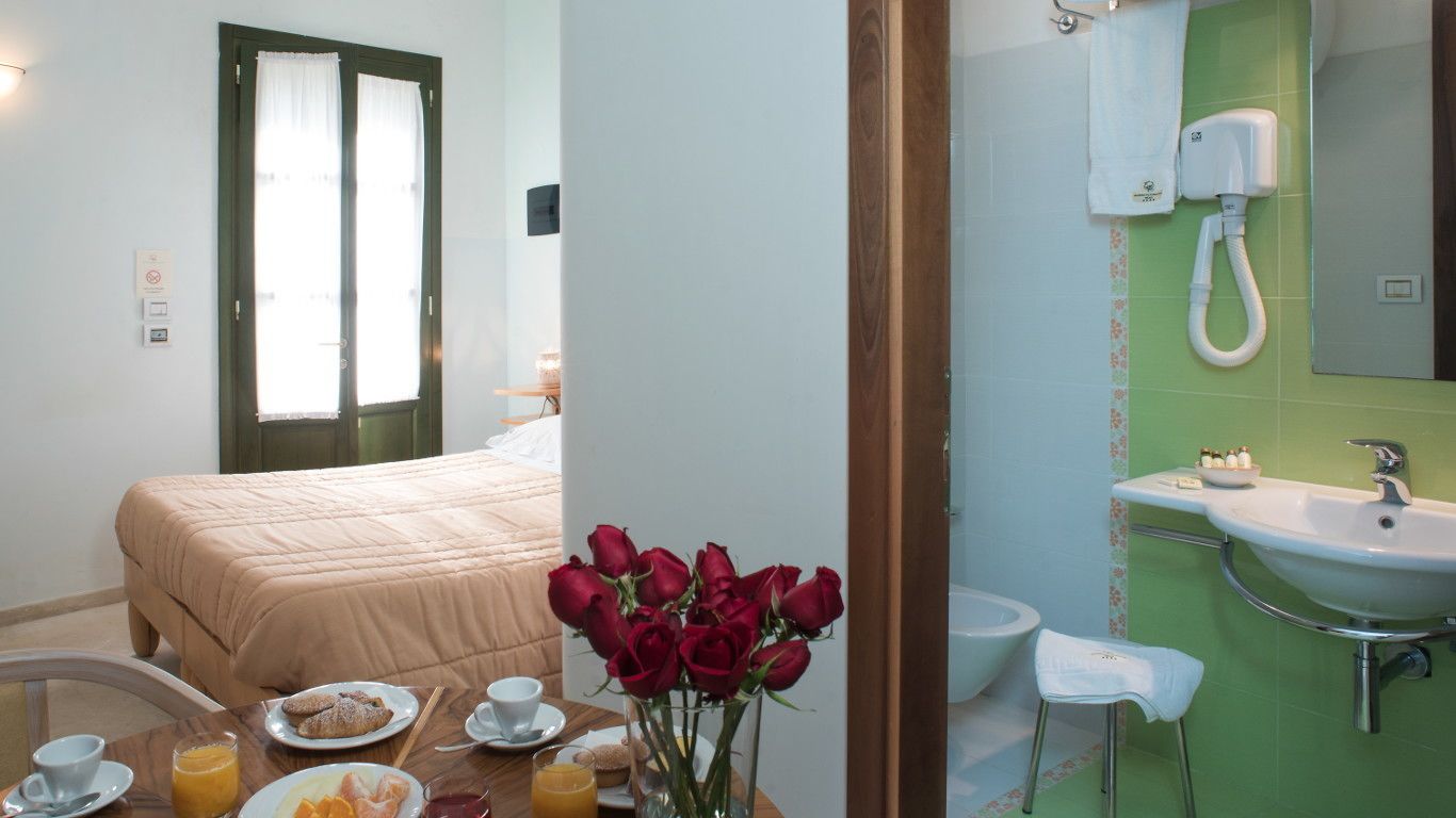 A hotel room with a bed, breakfast table with pastries and drinks, a vase of red roses, and a bathroom with a sink and mirror.