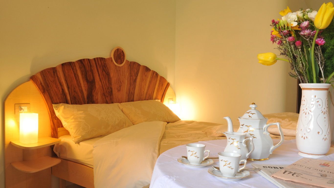 A neatly made bed with a wooden headboard, a bedside table with a lamp, and a table set with white tea service and a vase of flowers.