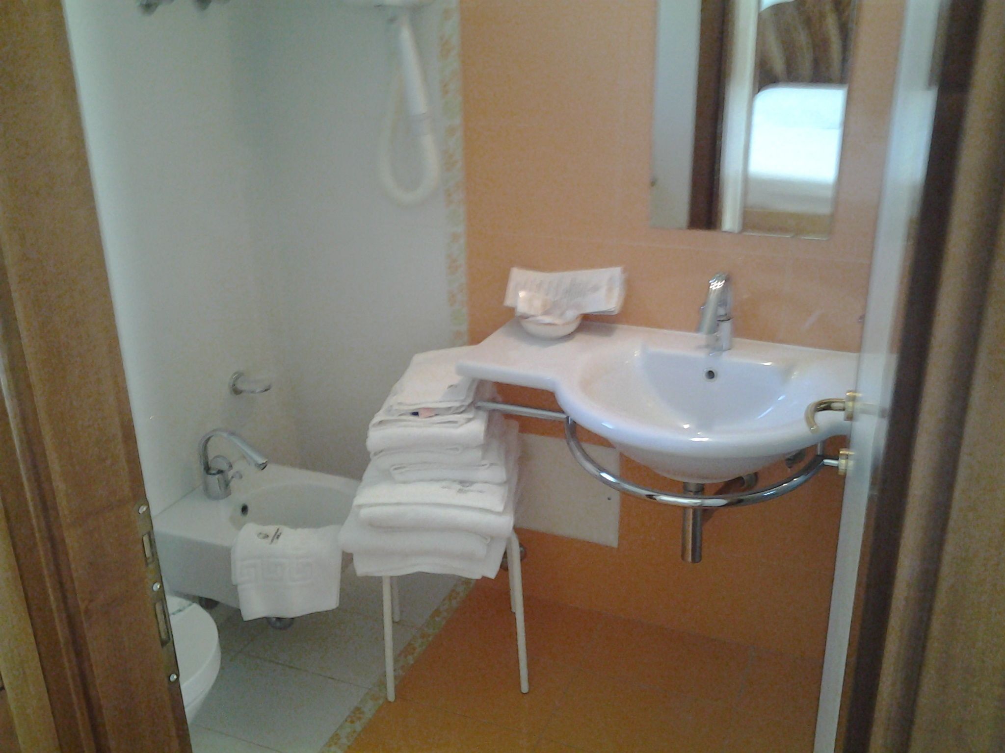 Ein Badezimmer mit einem weißen Waschbecken, einem Spiegel und einer Toilette. Handtücher liegen auf einem weißen Ständer gestapelt.