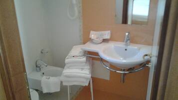 Ein Badezimmer mit einem weißen Waschbecken, einem Spiegel und einer Toilette. Handtücher liegen auf einem weißen Ständer gestapelt.