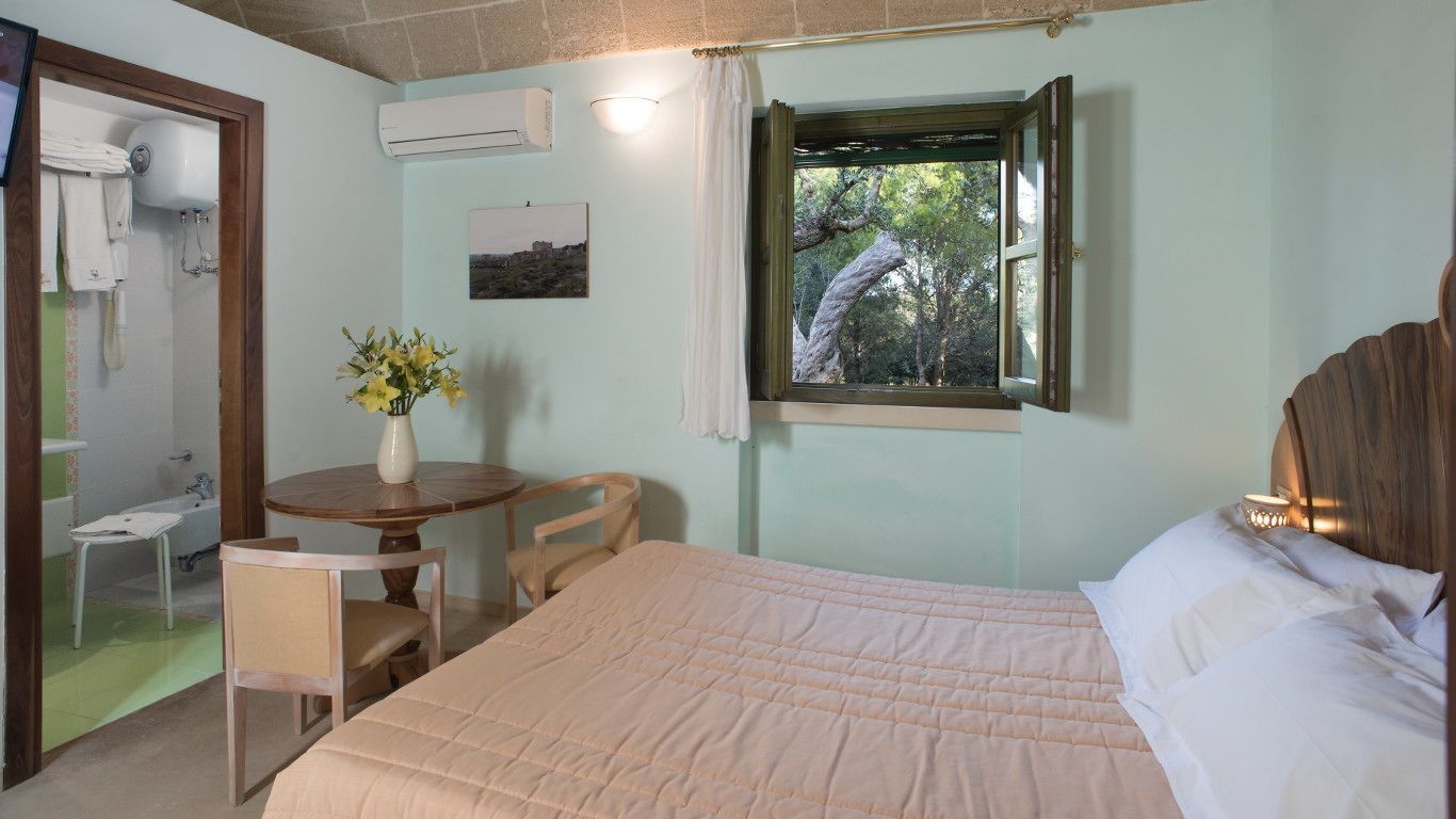 A bedroom with a bed, a bedside table with a vase of flowers, a window with a view of trees, and a bathroom visible through an open door.