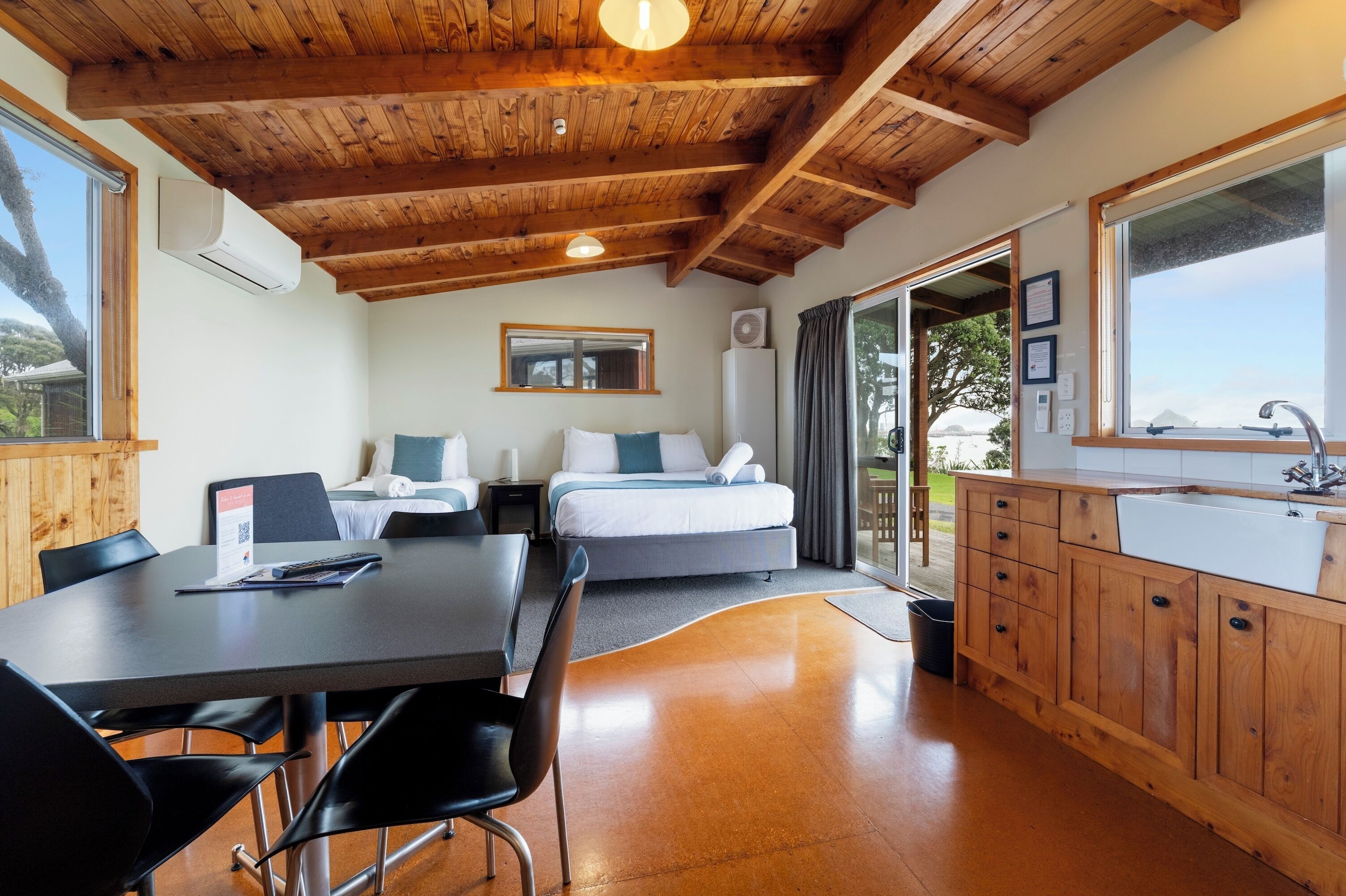 Quarto moderno com teto de madeira, uma janela grande, uma cama, uma mesa de jantar com cadeiras e uma cozinha com armários de madeira e pia.
